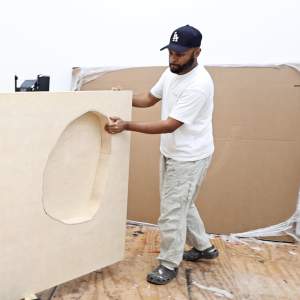 Shikeith rolls a large wooden shallow box sculpture across a white walled studio. He is a Black man with a dark beard, and wears a white tshirt and baseball hat.
