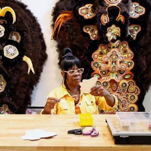 Denice Smith sits at a work table sewing pearl beads onto a patch. She is a Black woman wearing glasses and dark hair up in a bun. Behind her are two elaborate beaded regalias.
