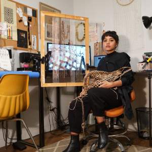The artist has medium-toned skin, and black bangs frame her face. She wears all black and holds a basket-like form in her lap. She sits in front of a frame strung vertically with beads, which is just in front of a desk with a computer and two sewing machines.