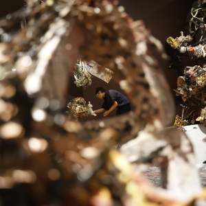 Seen through an opening in an out-of-focus abstract ceramic sculpture, Linda Sormin bends down to adjust an abstract sculpture of ceramic tangles. She wears dark clothing and glasses, and has short dark hair.