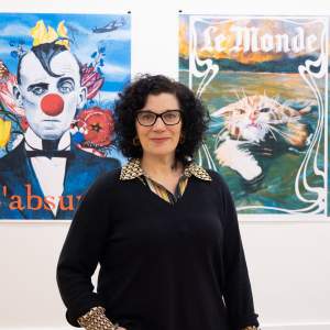 Laura Spector, a white woman with curly black hair and black frame glasses, stands facing the camera with a slight smile in front of two paintings.