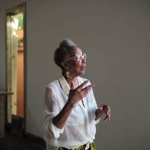 An Afro Puerto Rican woman with medium skin tone and short curly grey hair gestures with her hands while appearing to be speaking to someone outside of the photo. She is in a dimly lit artist room with a projected image in the background.