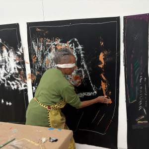 Awilda Sterling draws with pastels on black paper pinned to wall, blindfolded. She is an elder Afro-Rican woman with short grey hair and wearing a green sweater and apron.