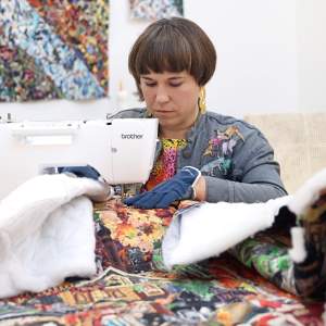 Jane Tardo, a white person with bobbed brown hair, works at a sewing machine on a large appliqué quilt. They wear earrings, a denim jacket and protective gloves.