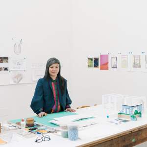 Lan Tuazon sits at a large studio table, which holds architectural models, and photos and drawings are pinned to the wall. She is a woman with Filipino heritage, and has long dark hair laced with gray and asymmetrical bangs. She wears a navy blazer and colorful button up underneath.