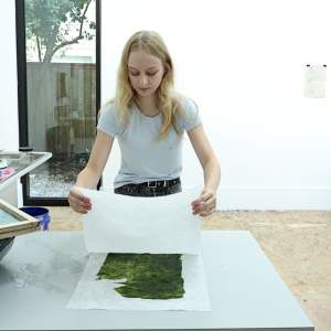 Katya Vaz, a white Russian American woman with light skin tone and light blonde hair, leans over paper in a white-walled studio.
