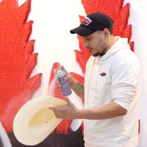 José sprays adhesive onto a cream colored cowboy hat. He is wearing a white hoodie and a black hat, and has medium light skin and dark facial hair.