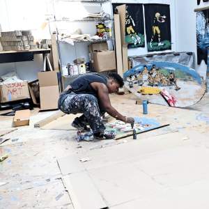 Bianca Walker, a Black person with short hair and paint-splattered clothing, crouches over cardboard, paints with their hands. They are surrounded by gestural figurative artwork on the walls and floor