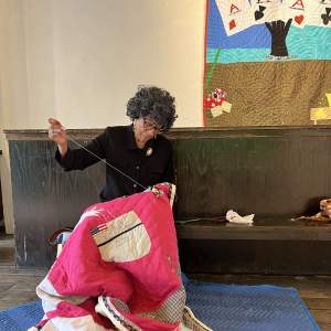 Yvonne Wells sits on a wooden bench, pulling thread up as she stitches a bright pink quilt with various shapes and colors. Yvonne is a Black woman with curly graying black hair and black-rimmed glasses. She wears a black dress shirt with a gold and pearl brooch and dangling earrings.