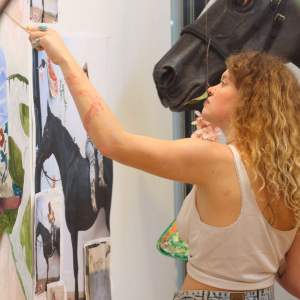 Summer's arm is extended, painting in front of a full size horse sculpture. She has light skin and long curly blonde hair.