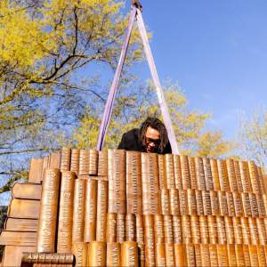 Sandy Williams IV leads over a sculpture of stacked books, hoisted by straps, amidst a backdrop of blue sky and green trees. Sandy is a Black person with medium-light skin tone and chin-length dreadlocks.