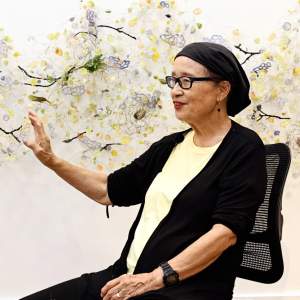 Yuriko smiles and gestures towards a sculptural installation on the wall. She is a senior Japanese-American woman, wearing a black hankercheif, glasses, and wears a black cardigan and light yellow shirt.