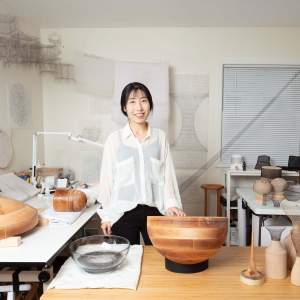 Artist Jayoung Yoon, a woman of Korean descent with light skin tone and black hair, stands in her studio—a room with off-white walls and several large work tables. We see multiple wooden molds and woven artworks on the surfaces of the tables, along with sketches on the walls.