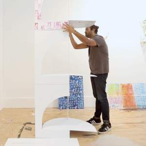 Artist Natan Diacon-Furtado arranges patterned and shaped foam core into a tower, surrounded by white walls with hanging cut paper artworks