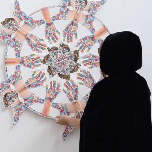 In a black hooded cape, artist Mary Grace Bernard hangs artwork of bejeweled hands and feet in a patterned circle.