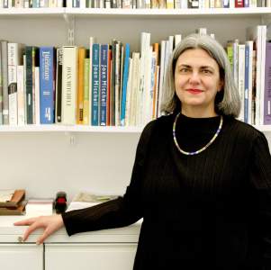 Alexandra Keiser stands in front of a bookshelf with various art catalogues. She is a white woman with shoulder length grey hair, wearing a black shirt and beaded necklace.