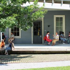 Pairs of people seated and talking to each other on a shaded porch of a blue-gray building with large windows, next to a tree and a grassy area.