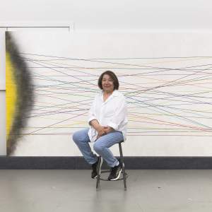 Virginia sits on a stool in front of a large, horizontal painting with intersecting colored lines in the center and areas of yellow fading to black on the sides. A Mexican-American woman in her mid eighties, Virginia has a brown bob and wears a white button-down shirt with jeans and black sneakers.
