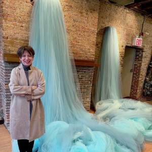 Ana María Hernando stands in front of two cascading waterfalls of blue tulle fabric in a brick walled room. She smiles with crossed arms, and is an Argentinian woman with dark short hair, wearing a trench coat and dark pants.