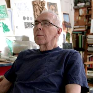An older white man with close cropped grey hair and glasses sits leaning back, small green linear paintings on paper hang in the background, next to a bookshelf