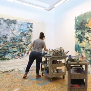 Standing in a white walled studio, a white woman with dark hair in a loose bun reaches for paintbrushes on a cart filled with painting supplies. On the walls are two large abstract paintings on unstretched canvas, filled with energetic gestural strokes creating tangles of movement.