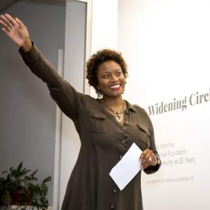 Shervone Neckles-Ortiz smiles with her right arm extended in the air. She is wearing an olive green buttoned dress and beaded pendant, and has medium dark skin and dark brown hair with tight curls.