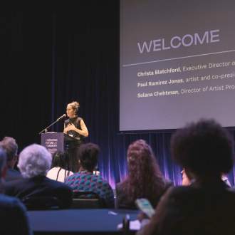 Solana Chehtman, a woman of Hispanic descent with light skin tone and brown hair pulled up in a bun, presents at a podium in front of a crowd seated at tables. On the screen behind her, we see "Welcome" along with her name and the names of other presenters.