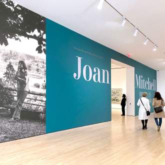 Two figures approach a large blue wall with text "Joan Mitchell" and large photograph of Joan Mitchell leaning on railing above the Seine River