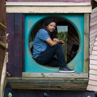Rontherin Ratliff, a Black man with medium skin tone and long black dreadlocks, sits in a round opening of an assemblage made from reused distressed housing materials