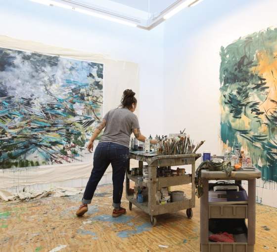 Standing in a white walled studio, a white woman with dark hair in a loose bun reaches for paintbrushes on a cart filled with painting supplies. On the walls are two large abstract paintings on unstretched canvas, filled with energetic gestural strokes creating tangles of movement.