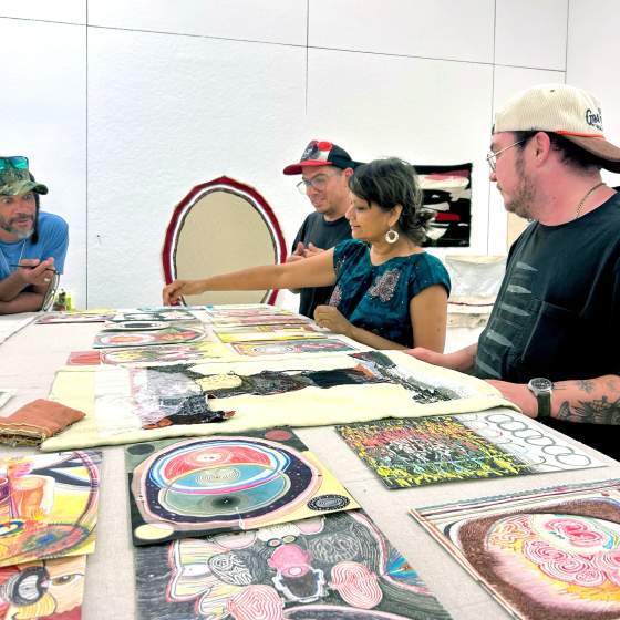 4 people of various age and appearance gather around a work table covered with colorful abstract artworks on paper and fabric..