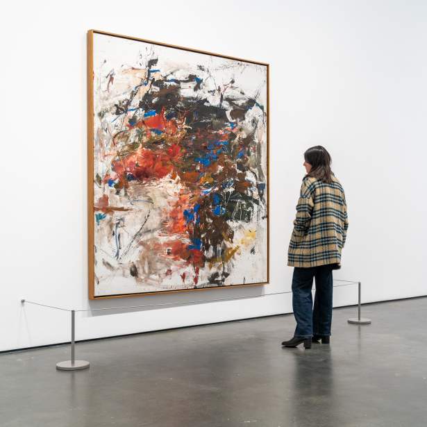In a white-walled gallery, a person with long brown hair and a plaid coat regards a large vertical abstract painting by Joan Mitchell. The painting is composed of bold intermixed strokes and daubs of brown, red, blue and green,creating a central form while leaving open areas of white ground around the perimeter, splattered and dripped with paint.