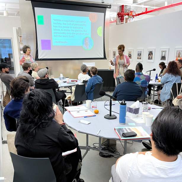 At a group of three round tables with white tablecloths, a group of about 16 people of various ages, dress, and skin tones look towards the front of the room, where two speakers stand: Noor Jones-Bey, a Black woman with medium-dark skin tone, clear glasses, and curly brown and blond hair, and Tiffany Lenoi-Jones, a Black woman with medium-light skin tone, bright green glasses, and black and gray curly hair.