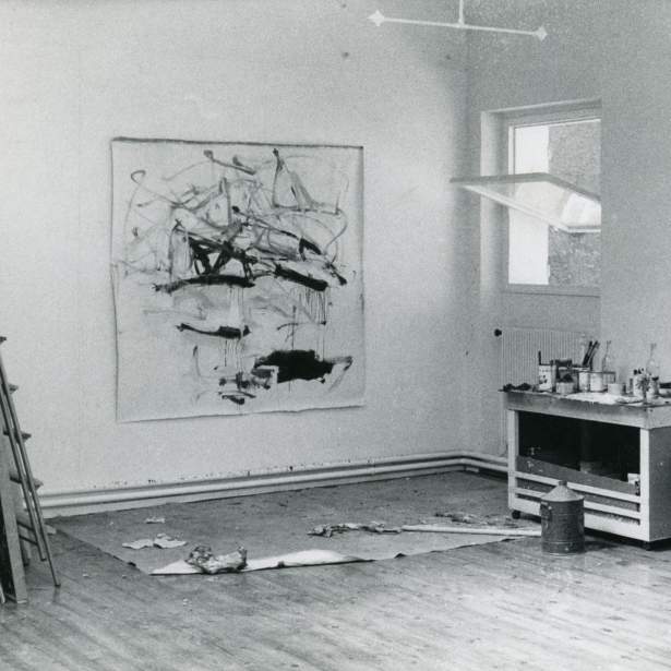 Black and white photo of art studio with large nearly square abstract painting on the back wall. To the right are two tables covered with art making supplies, and two large open casement windows.