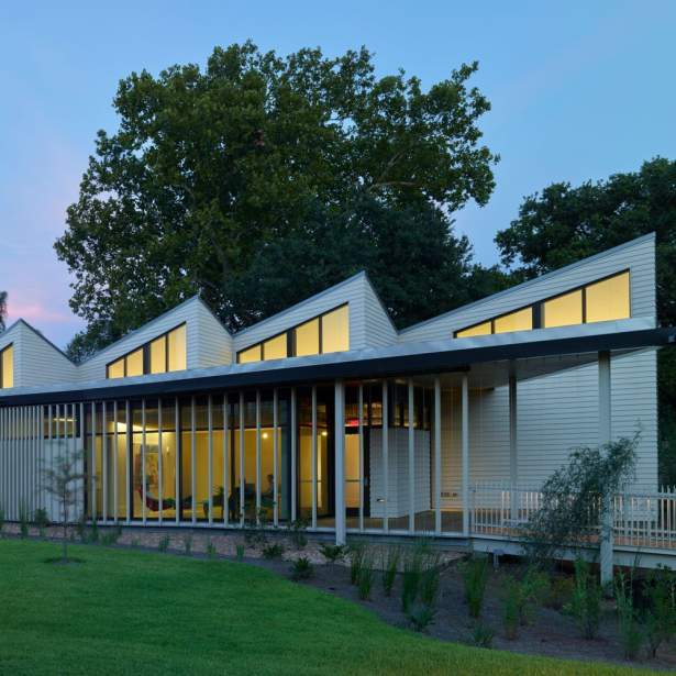 Studios at dusk - a modern siding-clad one-story building with four peaked studio roofs with warm light glowing from the skylights.