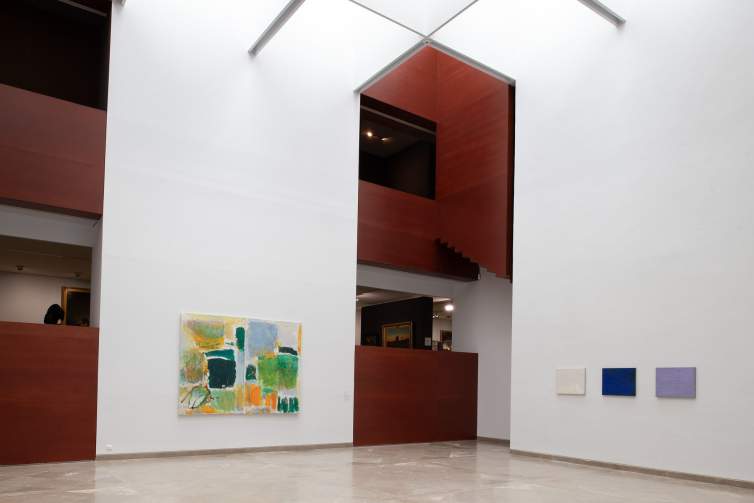 On white museum walls in an atrium with wood paneled architectural elements, four abstract artworks hang. The painting on the left wall is a diptych by Joan Mitchel with patches of vibrant colors in yellow, orange, blue, and green tones. On the right wall are three small paintings arranged in a horizontal row: one white, one cobalt blue, one lavender.