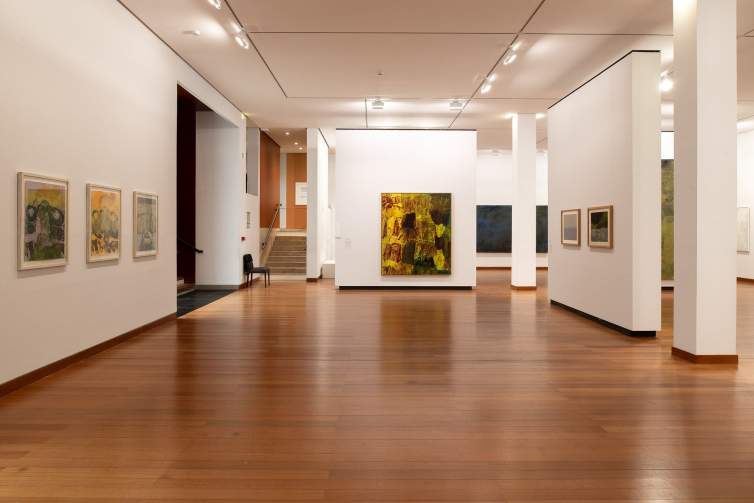 A variety of framed artworks hang in a museum gallery with white walls and columns and hardwood floors.