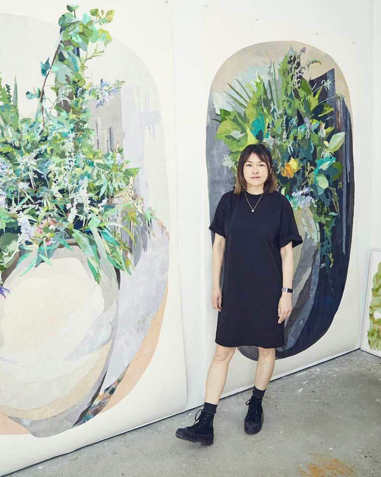 Naomi Kawanishi Reis stands in front of two large oval abstracted botanical paintings. She is a Japanese-American woman, wearing a black dress, and black boos, with mid length dark hair.