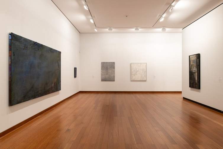 A variety of paintings hang in a museum gallery with white walls and hardwood floors.