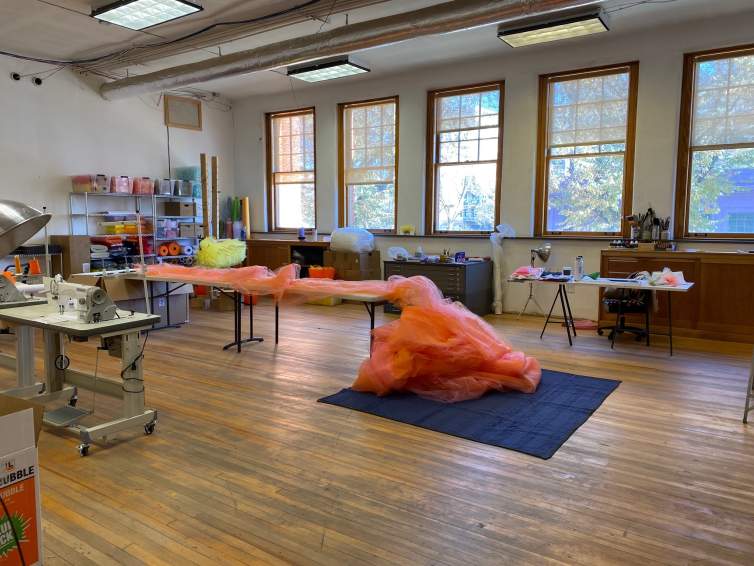 In a large room with wooden floors and wall of windows, artist Ana Maria Hernando’s tools and fabric artworks-in-progress can be seen on various shelves and tables. In the center of a room, a large pinkish-orange length of tulle fabric spills off a long table onto the floor.