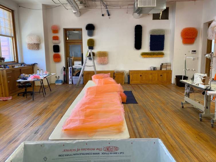 In a room with wooden floors and white walls dotted with abstract fabric sculpture, a long piece of pinkish orange tulle fabric lays on a central table. We see other work surfaces and tools in this artist’s studio.