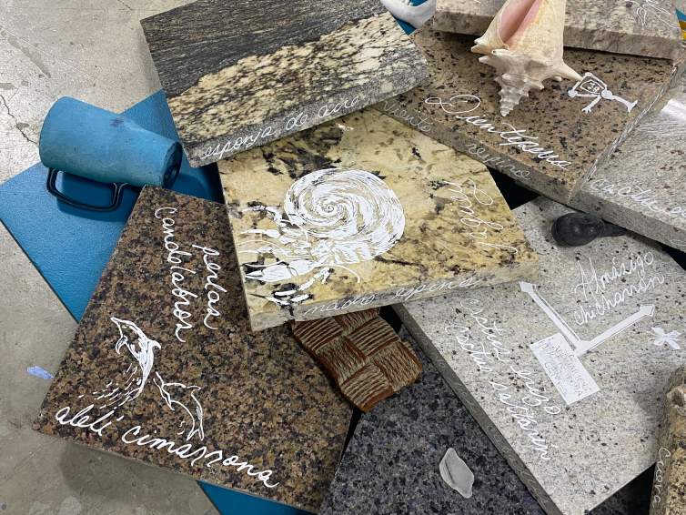 In a close-up photo of a sculptural installation, a pile of nearly a dozen square slabs of polished granite lay on a blue yoga mat with a travel cup, bones, seashells and other debris strewn amongst the slabs. The slabs have Spanish phrases and drawings on them, rendered in chalky white liquid paper, including drawings on shells, birds, and dolphins.