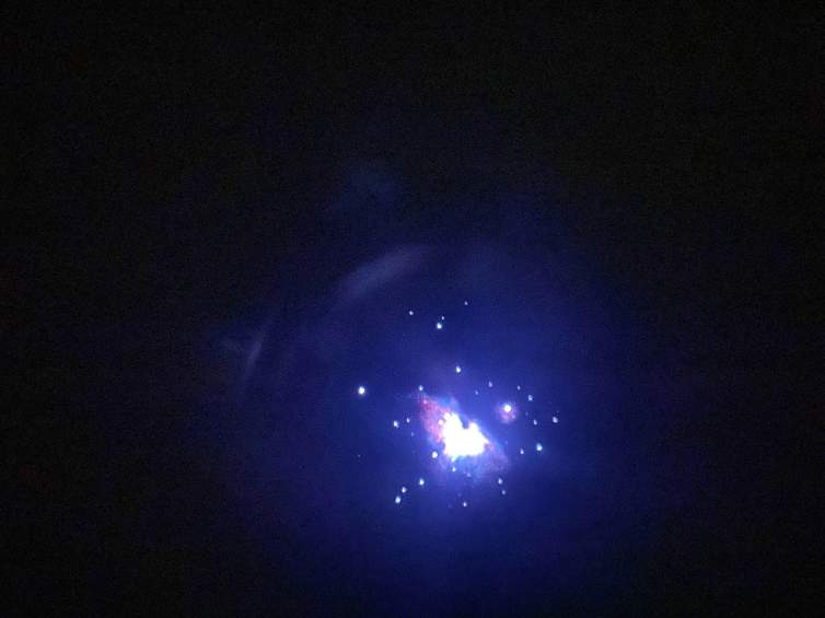 In a dark and blurry photograph of the sky, we see Orion’s Nebula captured with a telescope. It appears as a white heart shape amidst a halo of purple, dotted by white stars.