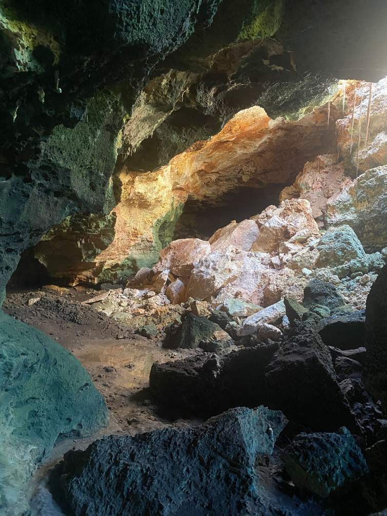A photo of the interior of a cave, with brown, gray, and white boulders strewn about and light entering the cave from the top right.