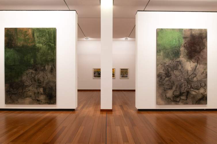 Two large abstract paintings hang in a museum gallery with white walls and columns and hardwood floors. We see three smaller works hanging on a wall further back in the gallery, behind a column.