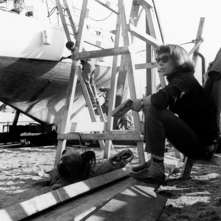 In a high-contrast black-and-white photo, Joan Mitchell sits on a scaffolding on a beach while a man works on a boat in the background. A white woman with chin-length hair, she holds a book and wears a black turtleneck, dark pants, penny loafers with socks, and sunglasses. A small dog sits at her feet.