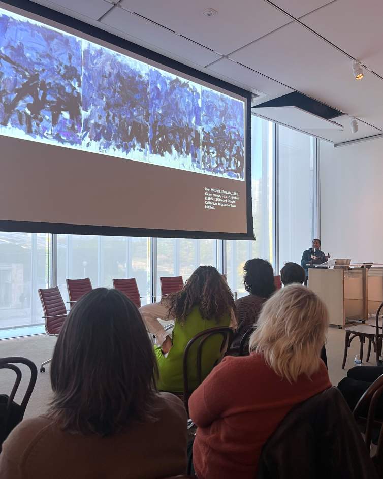 A large abstract painting is projected on a large screen in front of an audience. To the right of the screen, a Black woman with short dark hair at a podium looks towards the projection.
