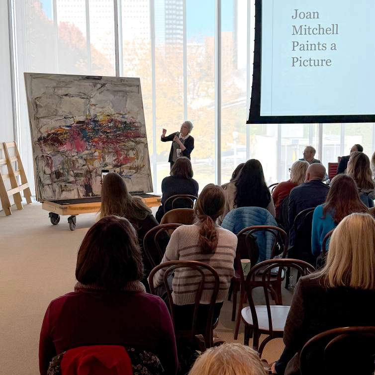 A large square abstract painting sits on a rolling dolly platform, as a white woman with grey hair, wearing glasses and dark clothing gestures towards a detail in the painting in front of a seated audience.