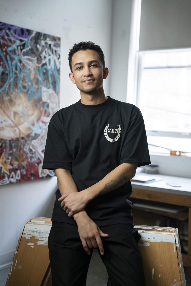 Anthony White leans on a saw horse in a studio in front of a painting and a window. He is a multi-racial person with medium skin tone, short curly dark hair and wears all black.