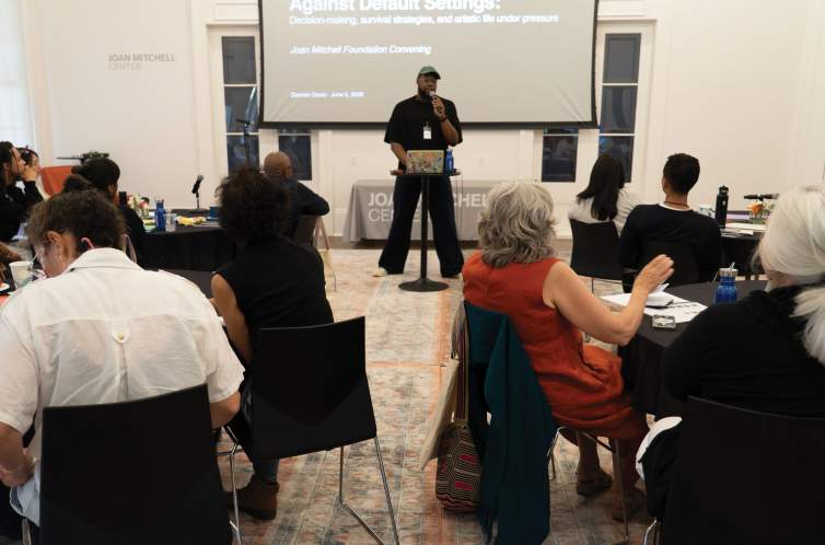 Damien Davis, a Black man wearing all black, glasses and a baseball hat, stands at the front of the room speaking into a microphone, in front of people gathered at round tables with black tablecloths.